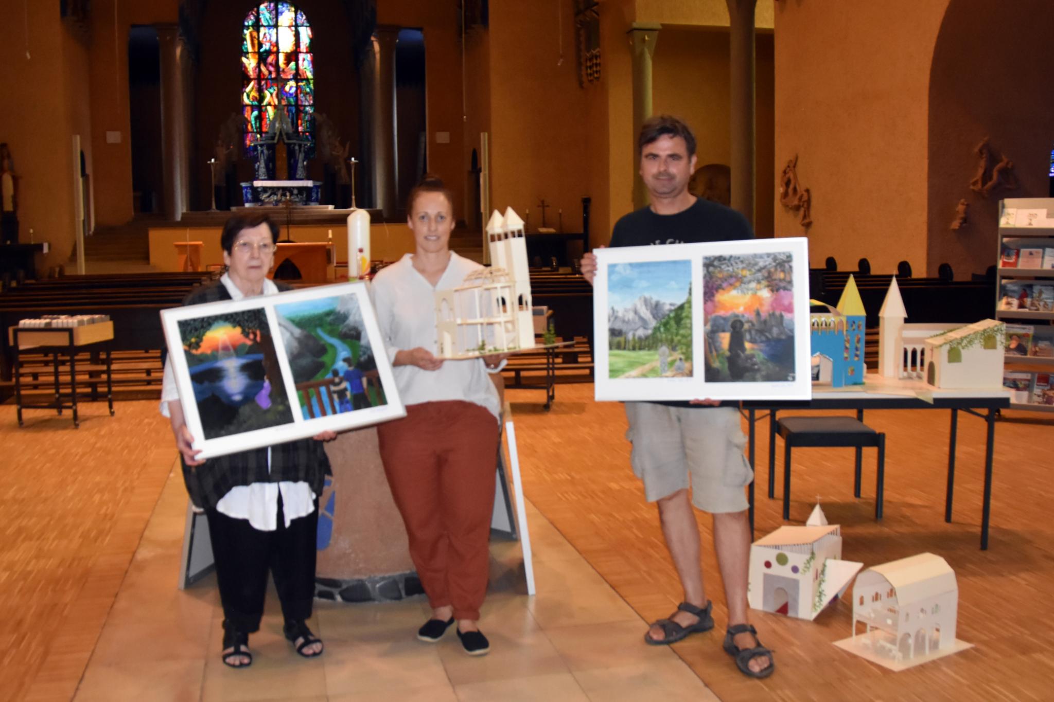 Man sieht drei Personen in einer Kirche stehen. Alle halten ein Kunstwerk der Schülerinnen und Schüler in der Hand.