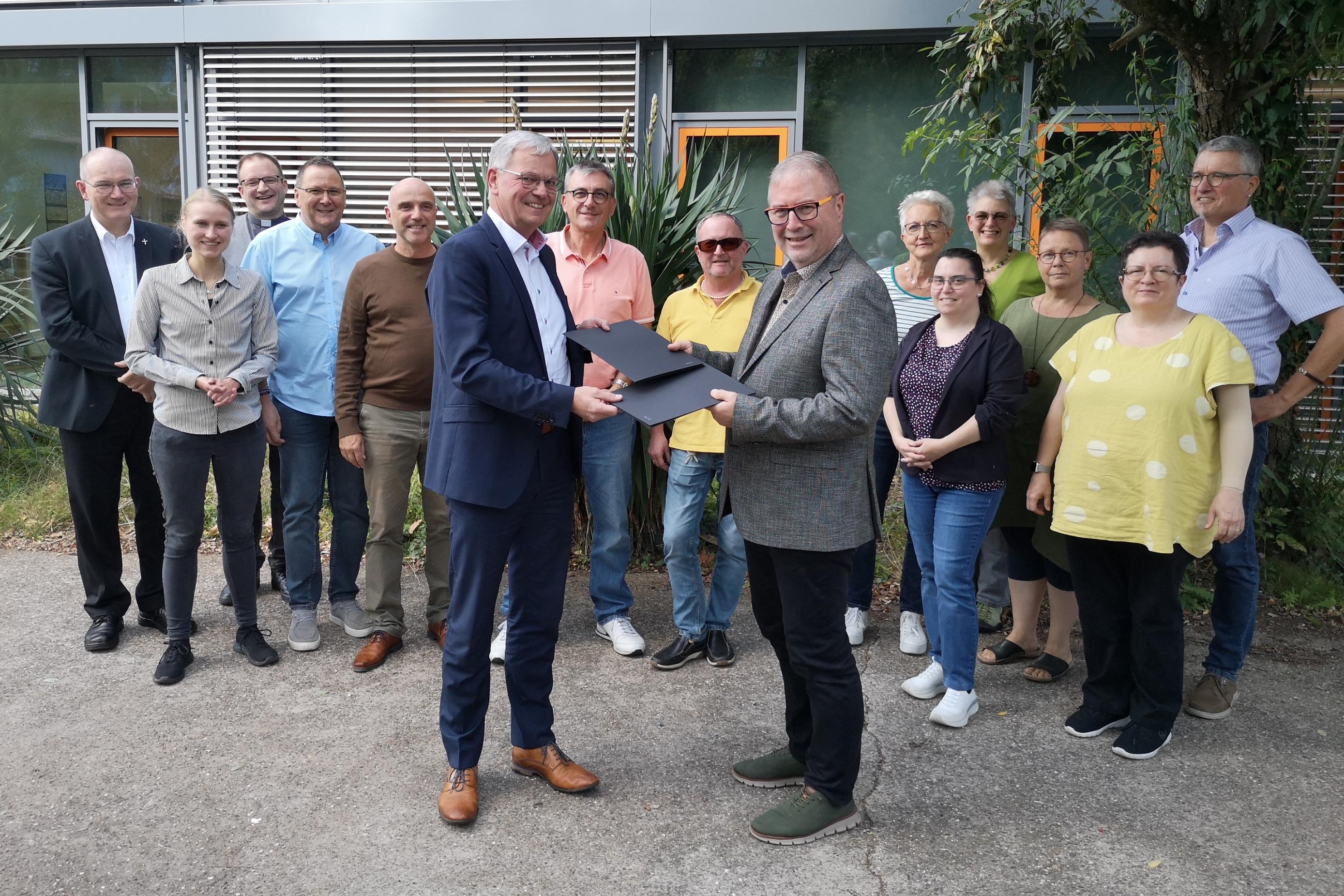 Generalvikar Dr. Ulrich Graf von Plettenberg (v.l.) und Superintendent Christian Weyer (v.r.) nehmen wechselseitig die unterzeichnete Kooperationsvereinbarung zur Krankenhausseelsorge in Empfang. Dahinter Vertretende der Klinikseelsorgenden im Saarland