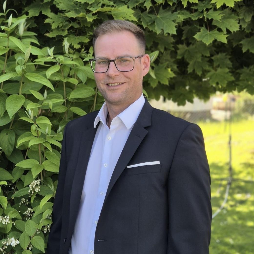 Ein Mann mit Brille und im Jacket steht vor einem grünen Busch und lächelt in die Kamera