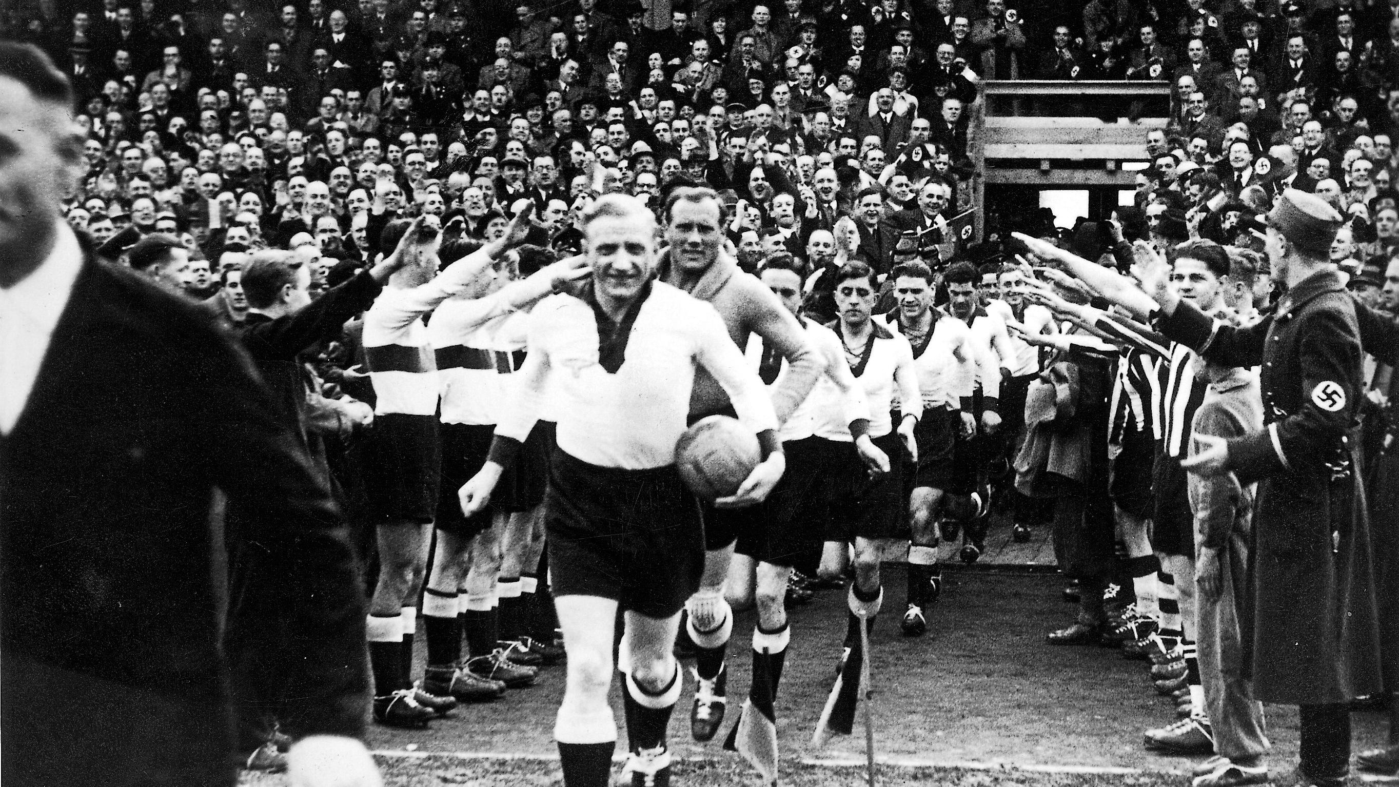 Historische Schwarz-Weiß-Fotografie eines Fußballstadions. Eine Reihe von Fußballspielern läuft aus einem Tunnel auf das Spielfeld. Die Person an der Spitze trägt einen Ball unter dem Arm und grüßt mit erhobener Hand. Links und rechts des Weges stehen zahlreiche Menschen dicht gedrängt, darunter auch uniformierte Personen; viele strecken den rechten Arm aus. Die Tribünen im Hintergrund sind bis auf den letzten Platz mit Zuschauern gefüllt. Die Aufnahme vermittelt den Eindruck eines großen Sportereignisses in einer politisch geprägten Atmosphäre der 1930er- oder 1940er-Jahre.