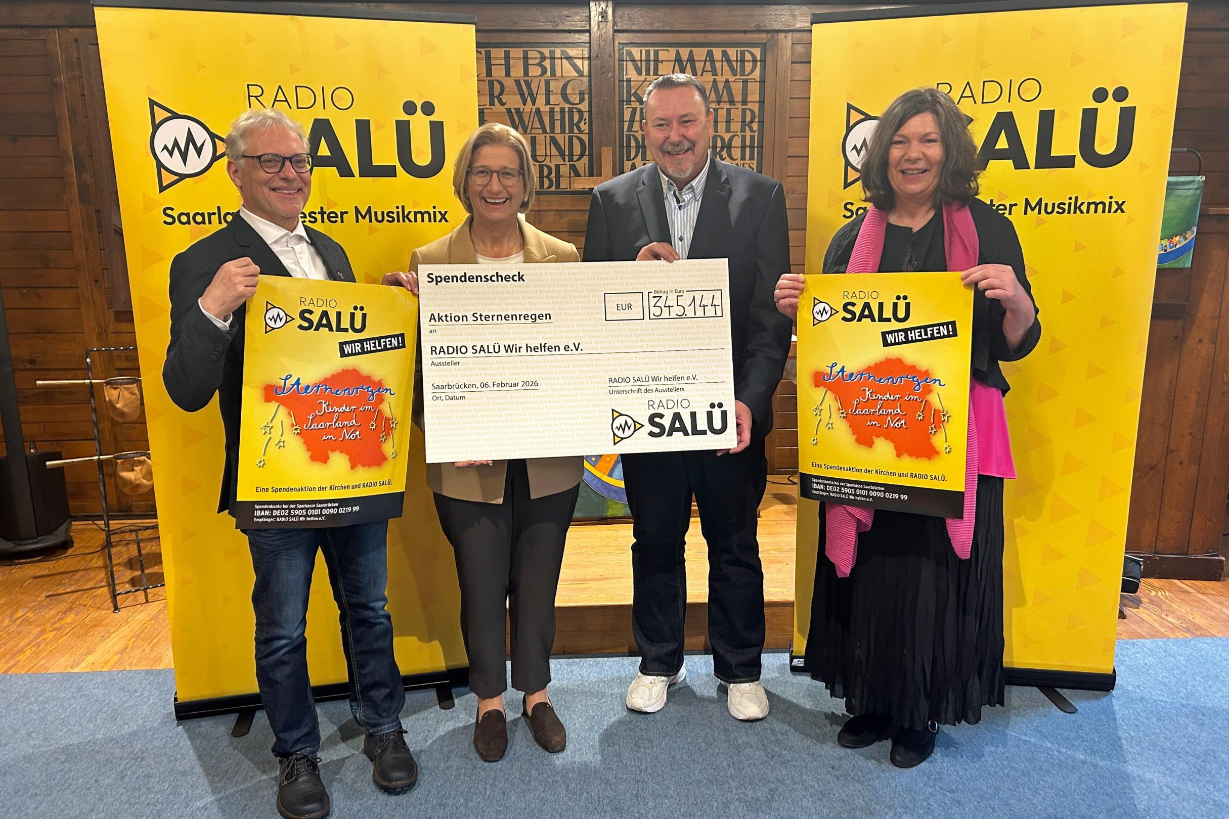 Vier Personen stehen in einem Innenraum vor zwei gelben Roll‑Up‑Bannern mit dem Logo von „Radio Salü“. Die beiden Personen in der Mitte halten gemeinsam einen großen Spendenscheck mit der Aufschrift „Aktion Sternenregen“ .Die beiden Personen außen halten jeweils ein farbiges Plakat derselben Spendenaktion. Der Boden besteht aus Holz, und im Hintergrund sind dunkle Holzwände mit eingravierten Schriftzügen zu sehen.
