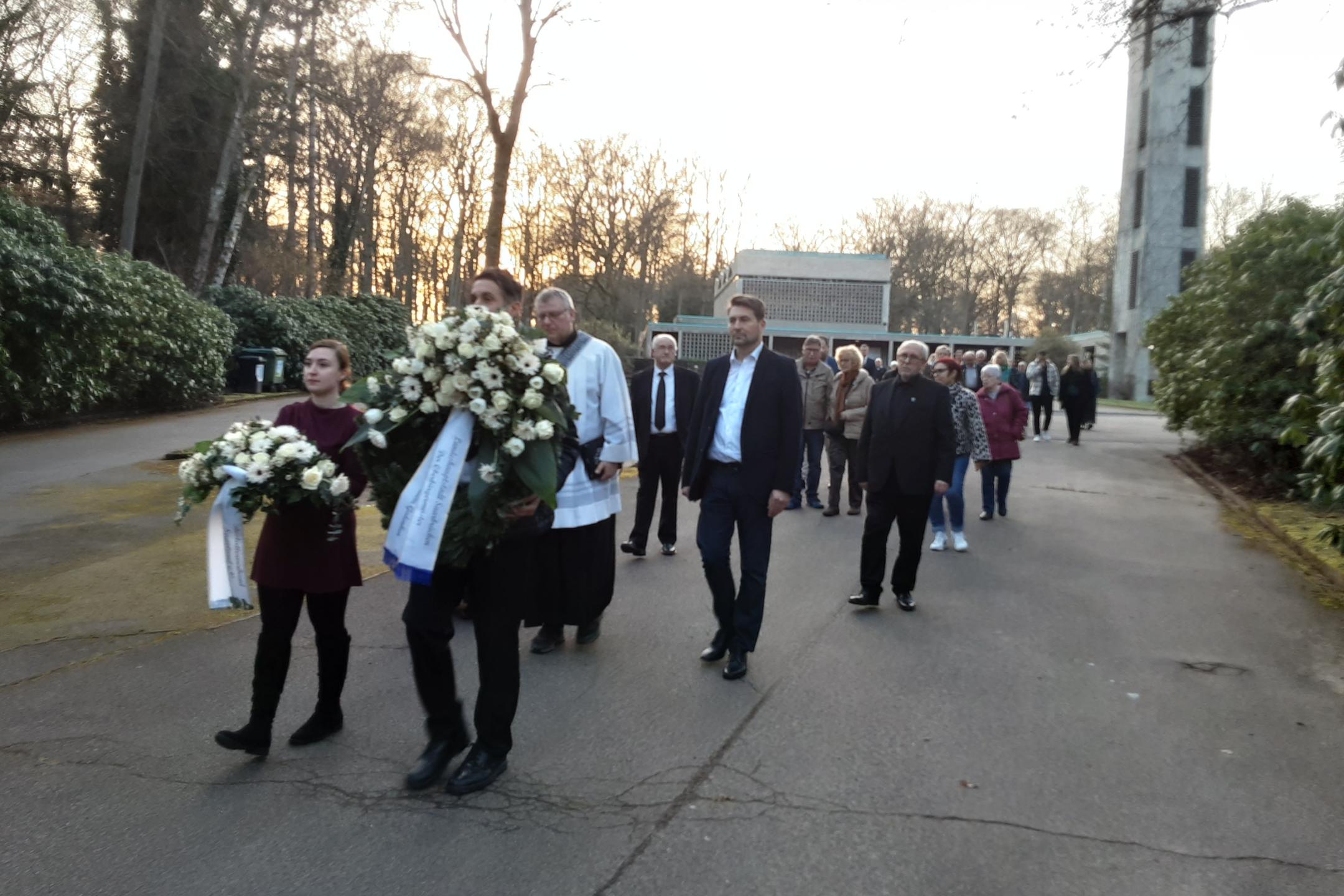 Eine Gruppe von Menschen geht in einem Trauerzug über einen breiten Friedhofsweg. Vorne tragen zwei Personen große weiße Kränze mit Trauerschleifen, begleitet von einer Person in liturgischer Kleidung. Dahinter folgen weitere Teilnehmende in respektvollem Abstand. Rechts im Hintergrund steht ein hoher Glockenturm, links und rechts säumen dichte Sträucher den Weg. Die Abendsonne scheint durch die kahlen Bäume und taucht die Szene in warmes Licht.
