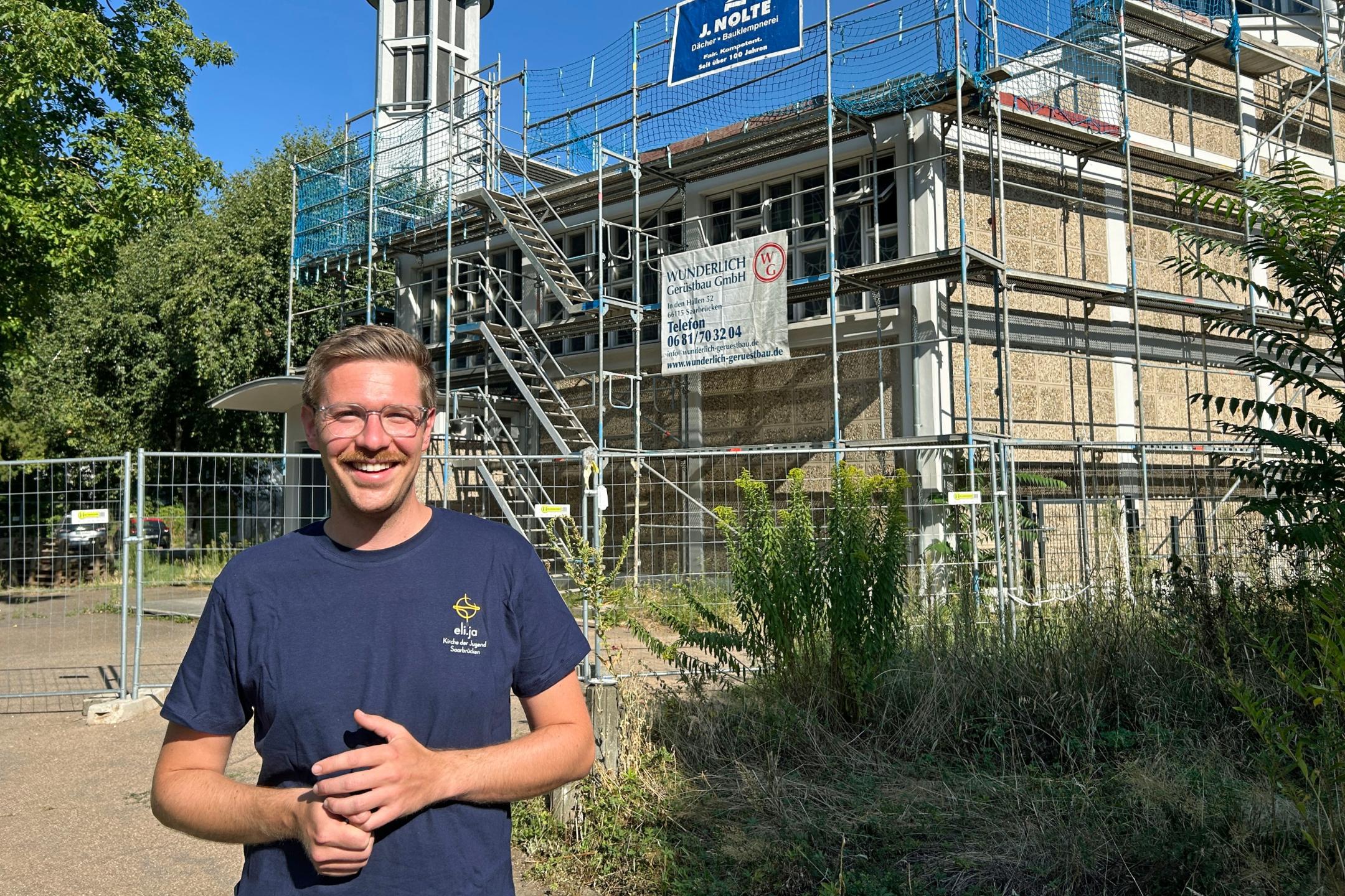 Ein Mann steht im Vordergrund auf einem Weg vor einem kirchlichen Gebäude. Das Gebäude ist vollständig von einem Gerüst mit Treppenaufgang umgeben und wird offenbar saniert. Hinter dem Gerüst ragt ein moderner Kirchturm mit rundem Dach und Kreuz in den blauen Himmel. Vor dem Gebäude stehen Bauzäune, am Gerüst hängen Werbebanner von Handwerksbetrieben. Links und rechts rahmen Bäume und Sträucher die Szene ein. Die Aufnahme entstand bei sonnigem Wetter mit klarem Himmel.