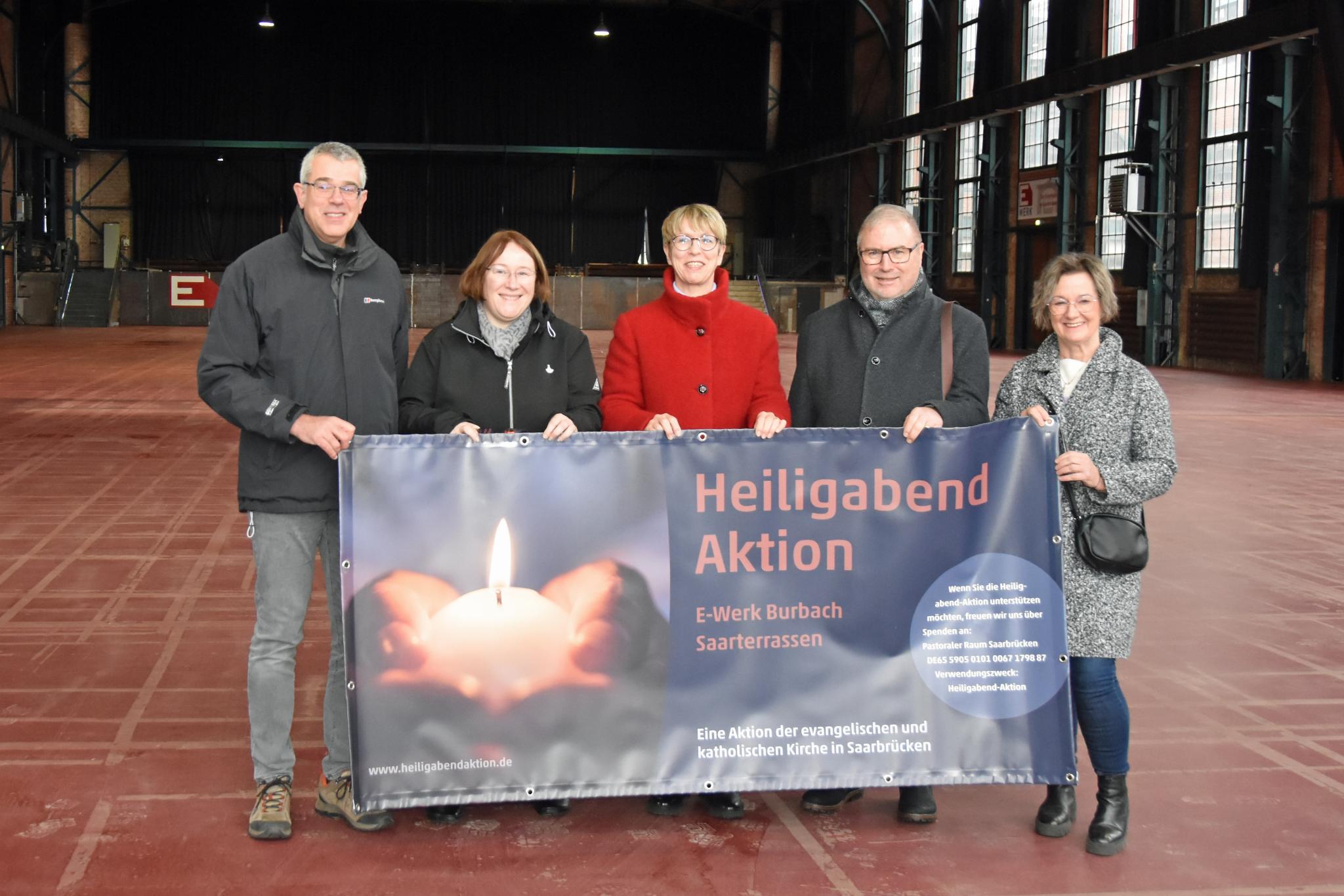 Fünf Personen stehen in einer Halle und halten das Plakat zur Heiligabend-Aktion.