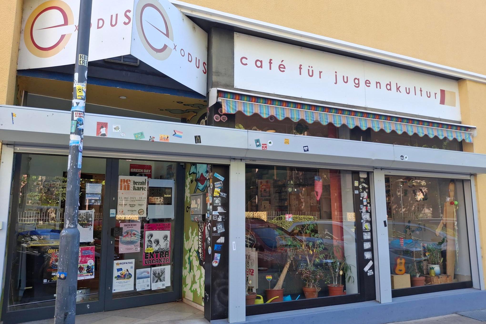 Eingang des ‚Café Exodus – Café für Jugendkultur‘ in einem gelben Gebäude. Über dem Eingang hängen zwei Schilder mit dem Schriftzug ‚Exodus‘ und ‚Café für Jugendkultur‘. Vor den großen Fenstern stehen viele Pflanzen, und die Glasflächen sind mit zahlreichen Stickern und Plakaten beklebt. Links befindet sich eine Glastür mit weiteren bunten Plakaten, die auf Veranstaltungen hinweisen. Ein Laternenpfahl steht am linken Bildrand und ist ebenfalls mit Stickern beklebt.