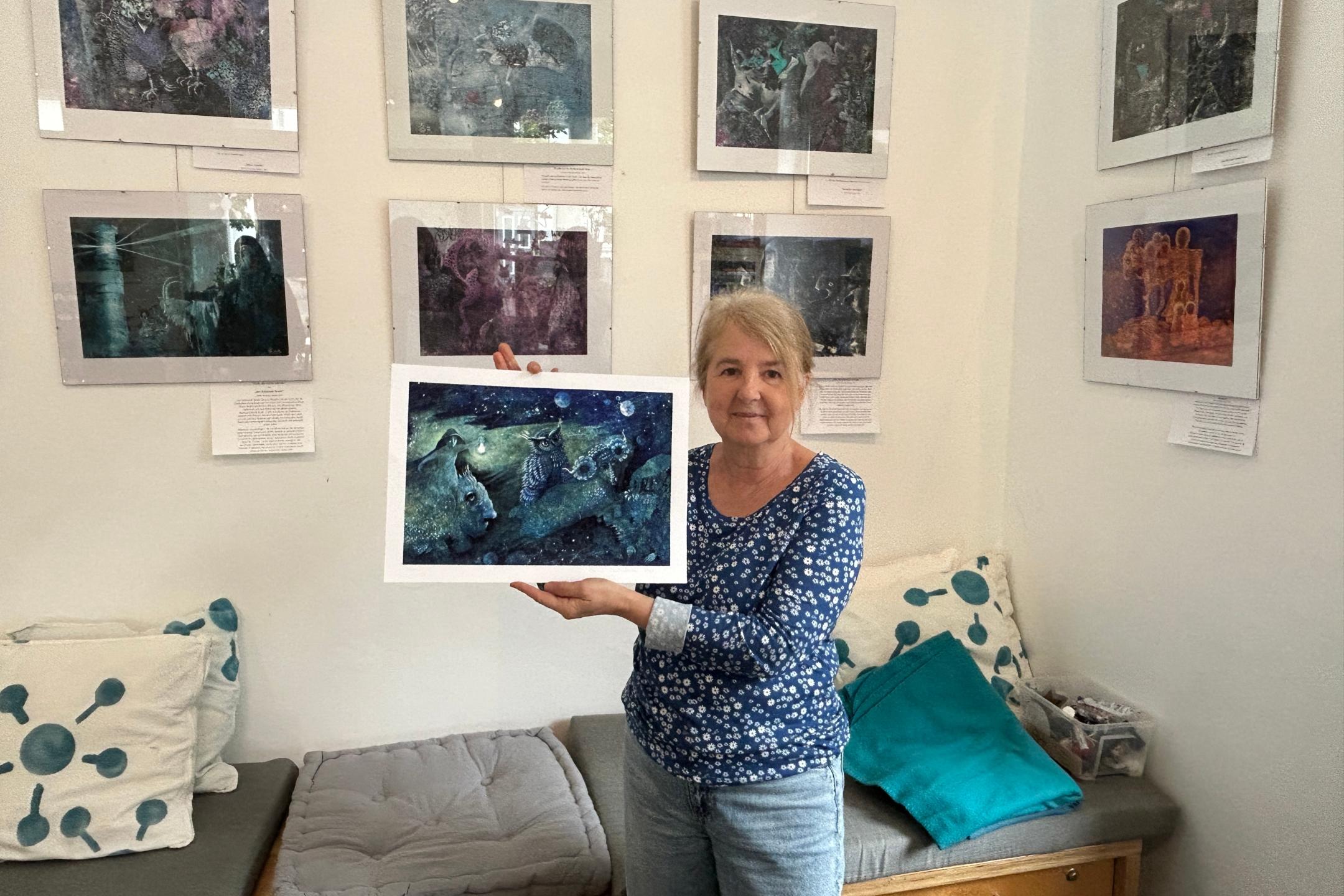 Eine Frau hält eins der von ihr gemalten Bilder in der Hand, weitere hängen im Hintergrund an der Wand.