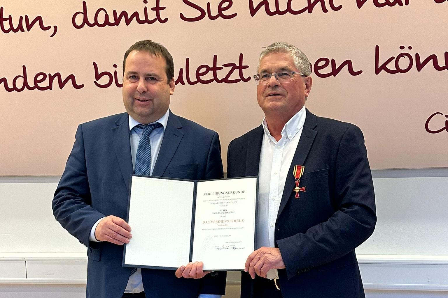Zwei Personen stehen nebeneinander in einem Innenraum vor einer großen Wand mit dekorativer Schrift in dunkler Farbe. Beide tragen formelle Kleidung. Die Person links (Sozialminister Magnus Jung) hält eine geöffnete Urkunde mit beiden Händen. Die Person rechts (Paul Herrlein) trägt ein Jackett, an dem ein Orden mit rotem Band befestigt ist, und berührt die Urkunde mit einer Hand. Der Boden ist aus hellem Holz, und hinter den beiden verläuft ein weißes Paneel entlang der Wand.